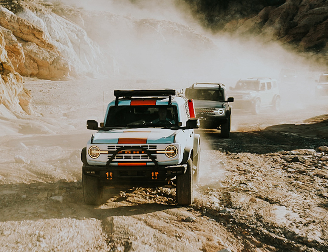 A bunch of broncos driving through a passageway kicking up dirt.